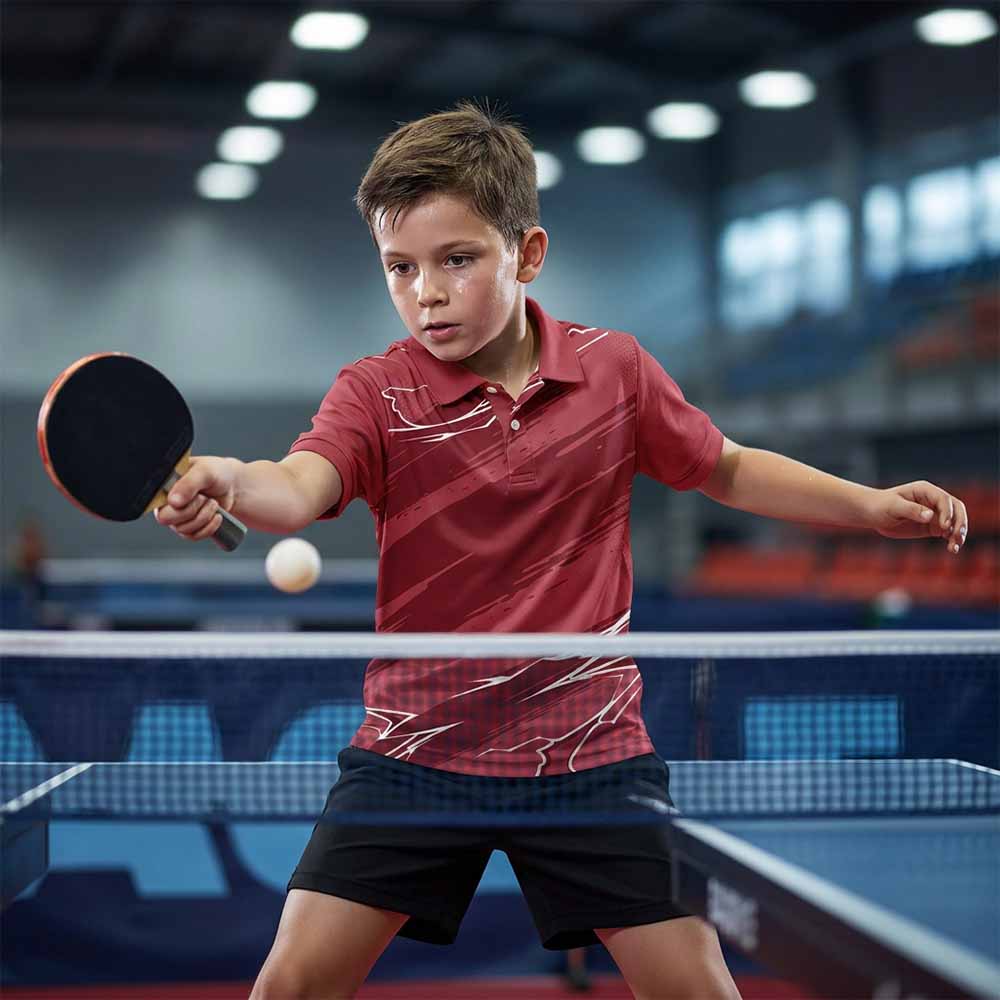 Kids' polo sports jersey in maroon with diagonal streaks and white angular accents, worn by a child in black shorts playing table tennis indoors.
