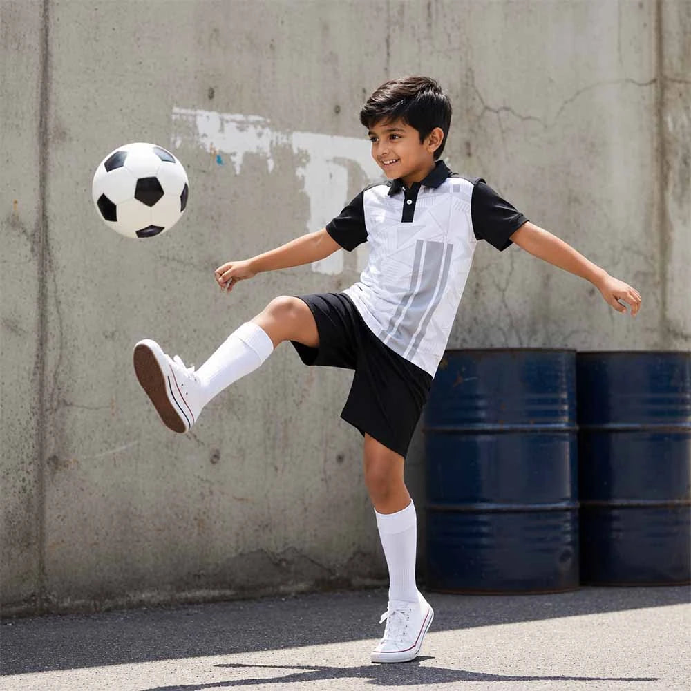 Child wearing a white and grey geometric graphics with black sleeves polo jersey and black shorts, kicking a football.