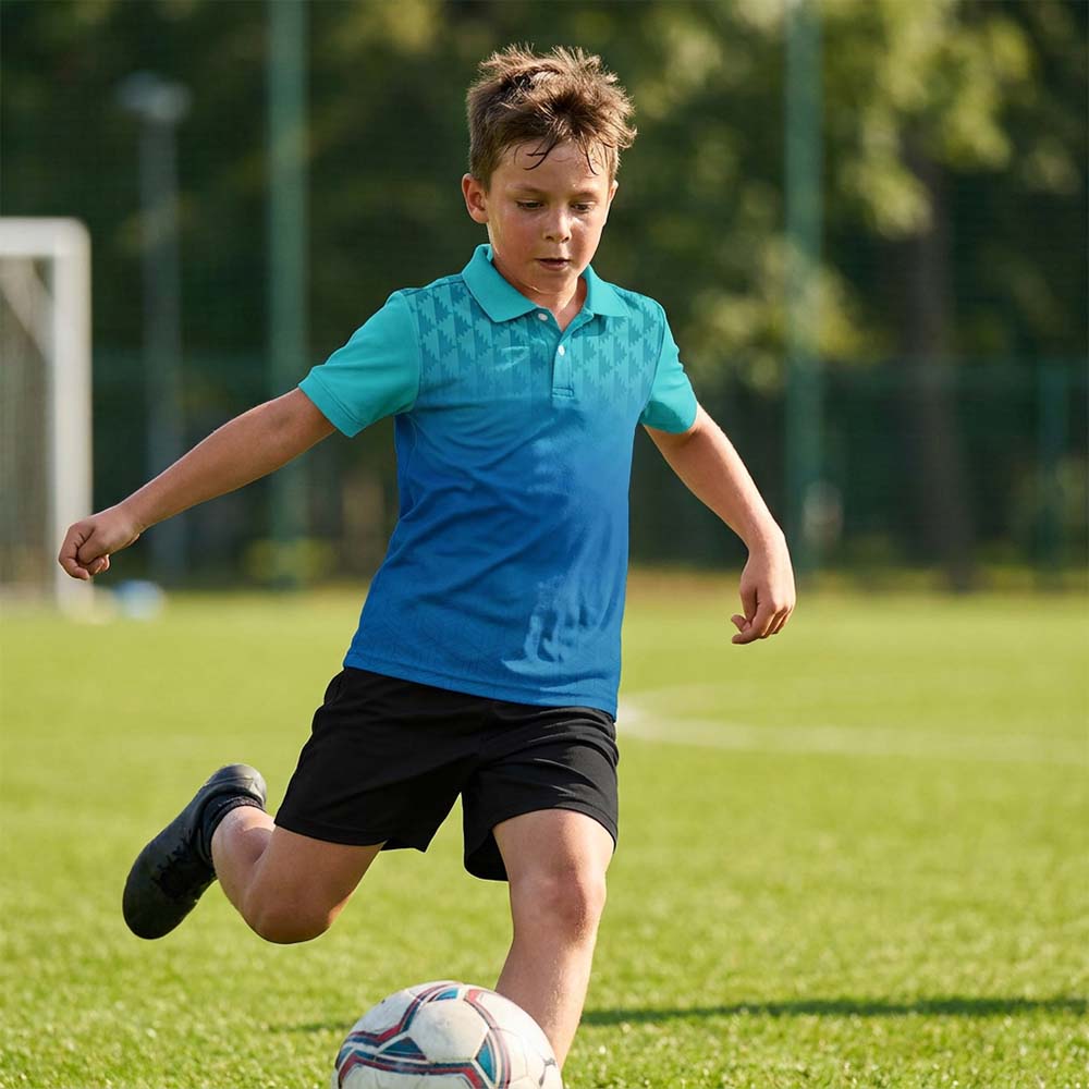 Kids' polo sports jersey in turquoise and blue gradient with a geometric shoulder pattern, worn by a child in black shorts kicking a football on grass.